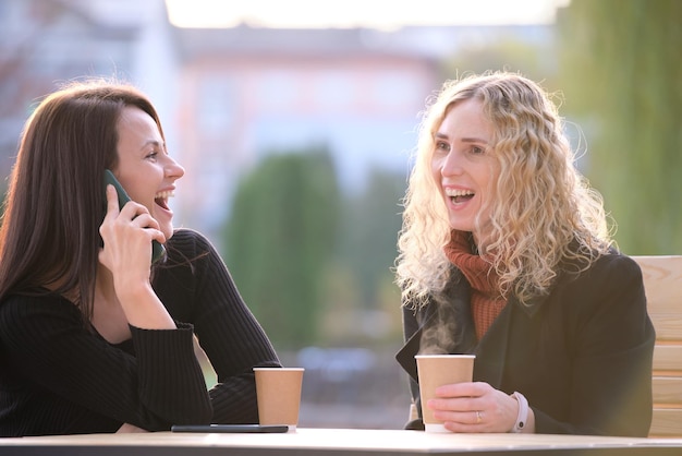 Fidanzate felici che si divertono insieme seduti al caffè di strada urbano ridendo e parlando al cellulare. Concetto di amicizia e attività all'aperto.