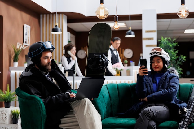 Fidanzato che utilizza il laptop e fidanzata con il telefono cellulare che gode della comunicazione digitale. Appassionati di sport invernali nella hall dell'hotel per divertirsi con lo sci e lo snowboard in attesa con i loro dispositivi intelligenti.