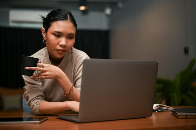 Fiduciosa donna d'affari asiatica che sorseggia caffè mentre guarda lo schermo del suo laptop