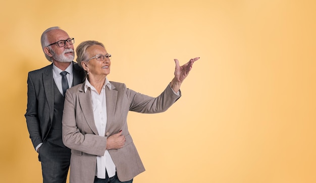 Fiduciosa donna d'affari senior che gesturing mostrando e indicando lo spazio della copia al dirigente professionista maschio in piedi con lei su sfondo arancione isolato