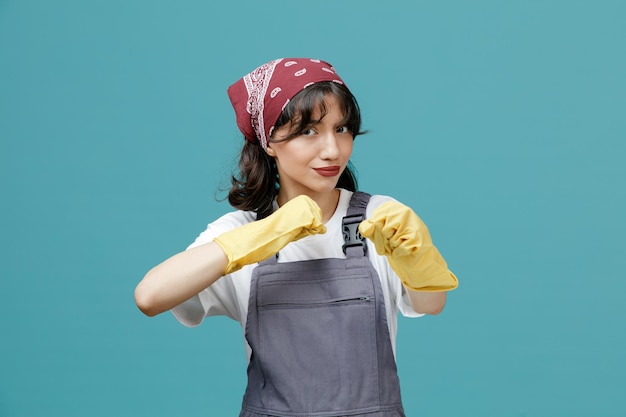 Fiduciosa giovane donna addetta alle pulizie che indossa bandana uniforme e guanti di gomma guardando la fotocamera che mostra il gesto di boxe isolato su sfondo blu