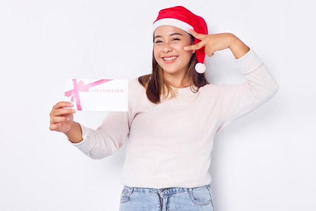 Fiduciosa giovane donna asiatica di 20 anni in un cappello di Babbo Natale che mostra un buono regalo coupon coupon card cover occhio con vittoria vsign gesto su sfondo bianco studio Buon Natale concetto