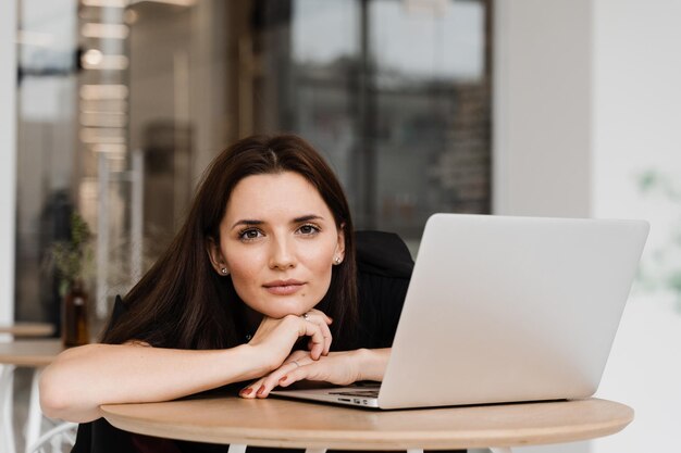 Fiduciosa giovane donna d'affari che lavora online con il team sul laptop sul progetto di avvio Attraente ragazza programmatrice con laptop sta lavorando online in un caffè bianco