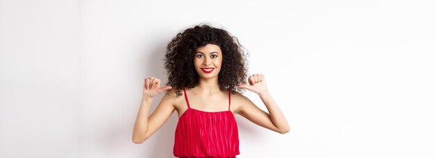 Fiduciosa giovane donna in elegante abito rosso che punta a se stessa e sorridente autopromozione in piedi ov