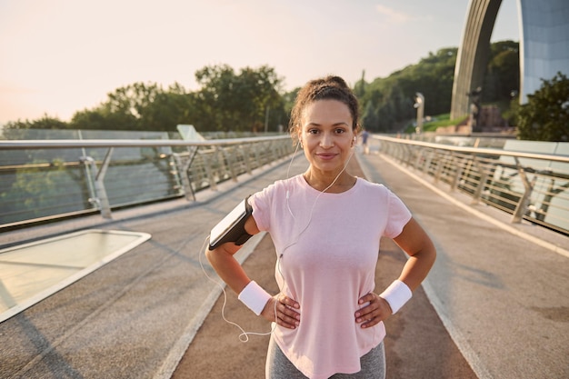 Fiduciosa sportiva ispanica, atleta femminile, corridore in maglietta rosa, indossa supporto per smartphone e auricolari, sorride guardando la fotocamera in piedi sul mulino del ponte della città, rilassante dopo la corsa