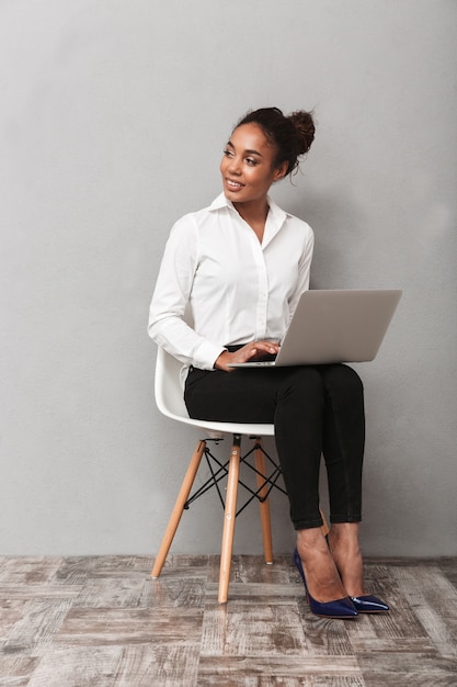 Fiducioso africano donna d'affari che indossa tuta seduto su una sedia, utilizzando il computer portatile su grigio