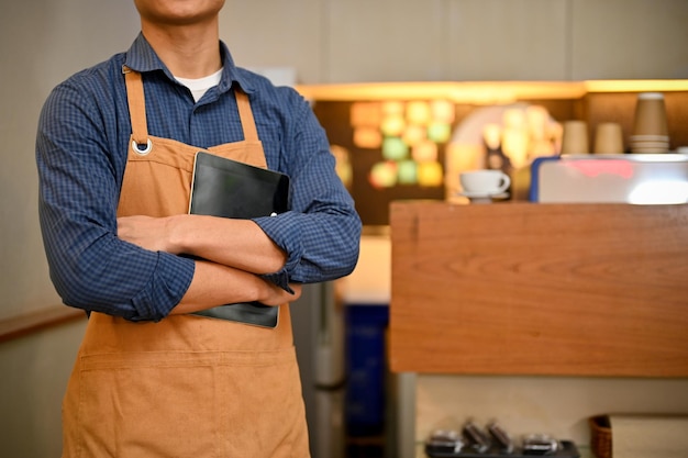 Fiducioso barista maschio asiatico o cameriere in grembiule sta con le braccia incrociate nella caffetteria