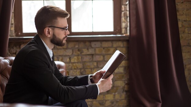 Fiducioso bell'uomo d'affari barbuto in abito elegante e occhiali che legge un libro o controlla i rapporti sul lavoro di sedersi sul divano in ufficio