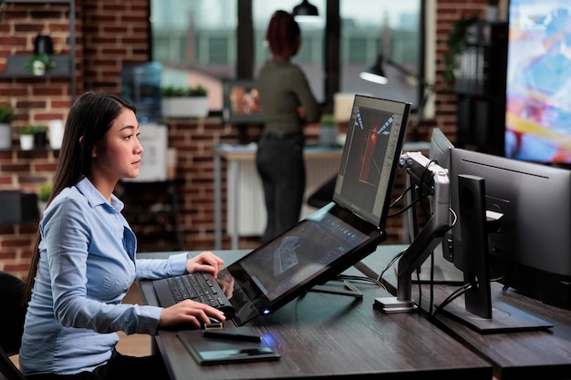 Fiducioso creatore di modelli 3D asiatico seduto alla scrivania con più monitor mentre si lavora su CGI. Artista digitale che crea immagini generate al computer per lo sviluppo di giochi mentre si trova nell'area di lavoro dell'agenzia creativa