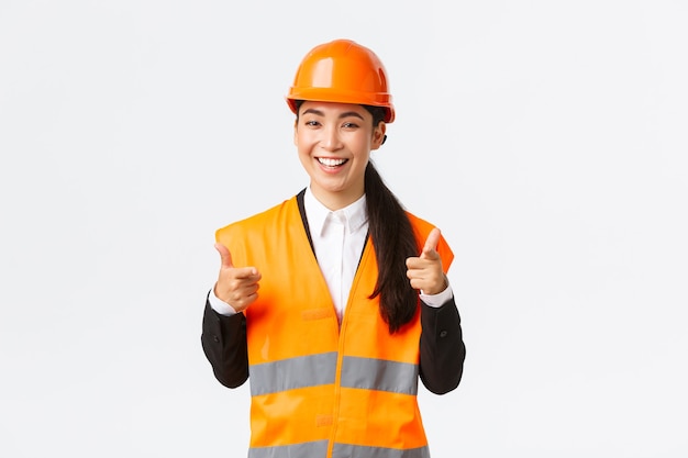 Fiducioso direttore di costruzione femminile piuttosto asiatico in casco di sicurezza e giacca riflettente, puntando il dito verso la telecamera lodando una buona scelta, un buon lavoro o ben fatto, in piedi sfondo bianco