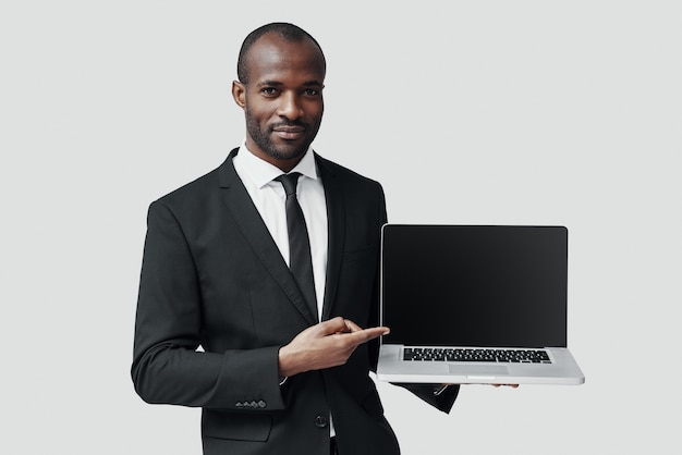 Fiducioso giovane africano in abbigliamento formale che indica lo spazio della copia sul computer portatile e sorride mentre sta in piedi contro il muro grigio