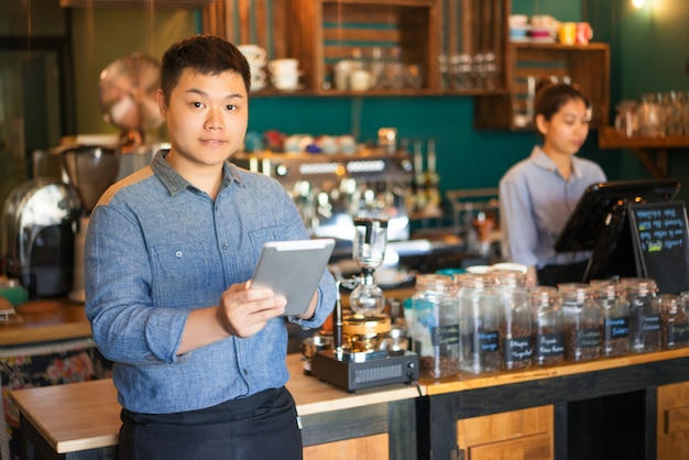 fiducioso giovane cameriere maschio asiatico in piedi al bancone e utilizzando il touchpad in caffetteria