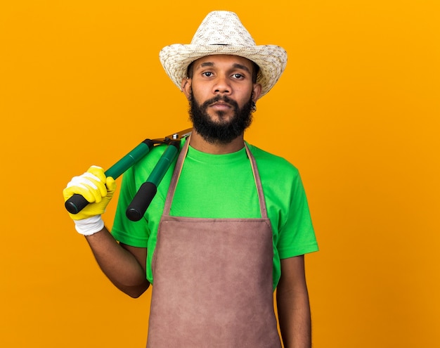 Fiducioso giovane giardiniere afro-americano che indossa cappello e guanti da giardinaggio che mettono le forbici sulla spalla