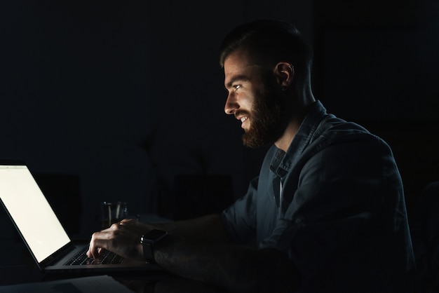 Fiducioso giovane sorridente che lavora al computer portatile mentre è seduto al tavolo al chiuso di notte