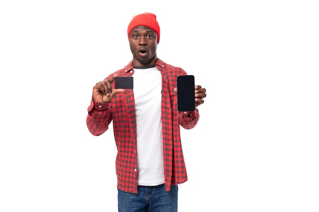 Fiducioso giovane uomo americano in copricapo rosso e camicia che mostra con carta mockup mano e smartphone
