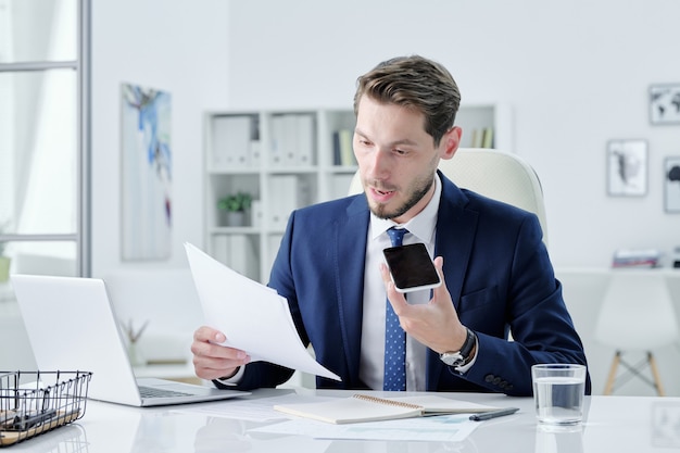 Fiducioso giovane uomo d'affari barbuto in abito blu scuro seduto alla scrivania e la registrazione di un messaggio vocale per il collega mentre commenta il contratto