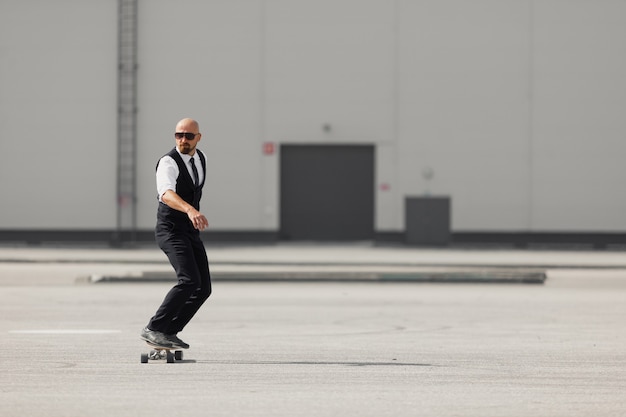 Fiducioso giovane uomo d'affari in occhiali che camminano per strada, utilizzando longboard.