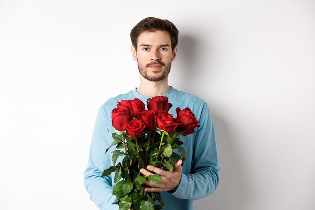 Fiducioso giovane uomo portare fiori il giorno di San Valentino, tenendo romantico bouquet, in piedi su sfondo bianco.