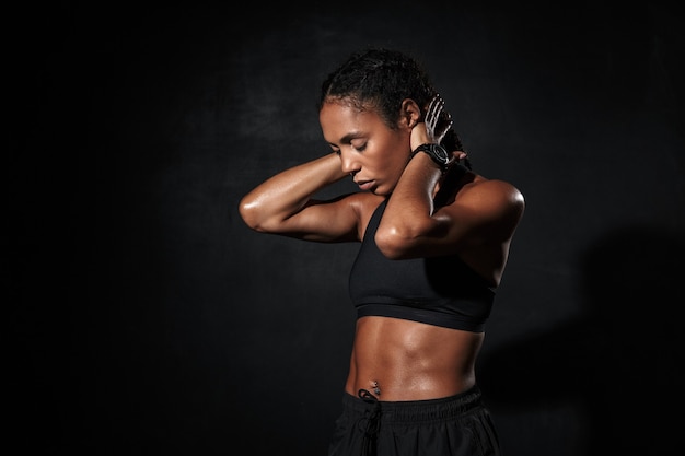 Fiducioso in forma giovane donna fitness africana che fa esercizi di stretching mentre si sta in piedi isolata sul nero