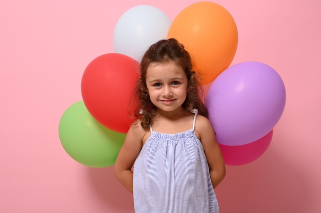 Fiducioso ritratto della splendida piccola ragazza di compleanno 4 anni con palloncini multicolori dietro la schiena e sorridente guardando la telecamera in posa su sfondo rosa con spazio di copia