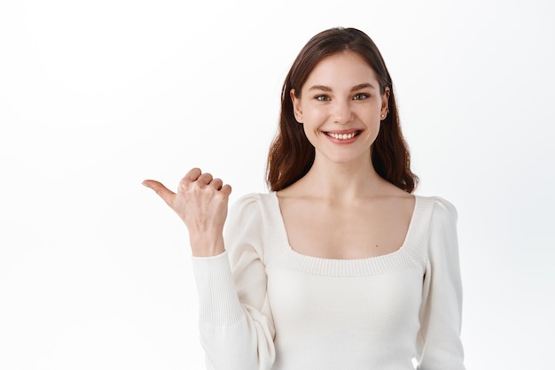 Fiducioso studentessa giovane professionista sorridente a te e puntando a sinistra da parte mostrando logo banner copypsace per il testo della tua azienda in piedi su sfondo bianco