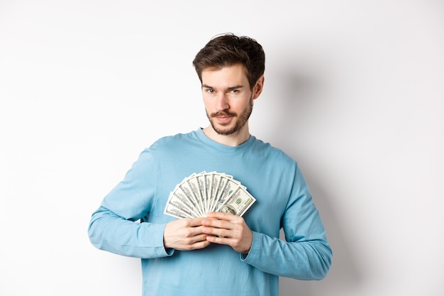 Fiducioso uomo barbuto sorridente, mostrando dollari e guardando la fotocamera. Bel ragazzo che tiene soldi su sfondo bianco