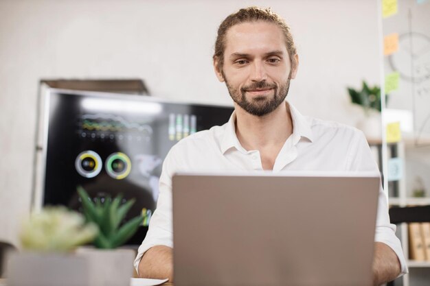 Fiducioso uomo caucasico in camicia bianca seduto in un ufficio moderno e utilizzando un computer portatile wireless