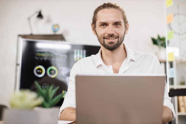 Fiducioso uomo caucasico in camicia bianca seduto in un ufficio moderno e utilizzando un computer portatile wireless