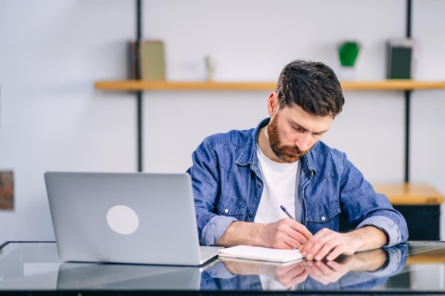 Fiducioso uomo d'affari che scrive note o relazione finanziaria seduto al tavolo con il computer portatile