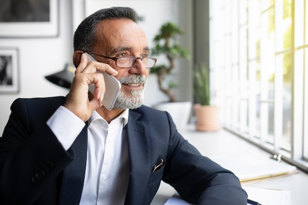 Fiducioso uomo d'affari europeo senior in giacca e occhiali chiama per telefono al tavolo ha una riunione a distanza