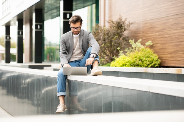 Fiducioso uomo d'affari in abbigliamento casual intelligente seduto davanti al laptop durante il networking o la chat video in ambiente urbano