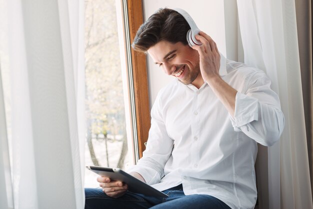 fiducioso uomo felice che indossa una camicia bianca utilizzando computer tablet e cuffie mentre era seduto alla finestra nel soggiorno