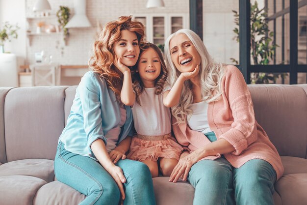 Figlia carina seduta sul divano con madre e nonna e toccando i loro volti con le mani e guardando la telecamera