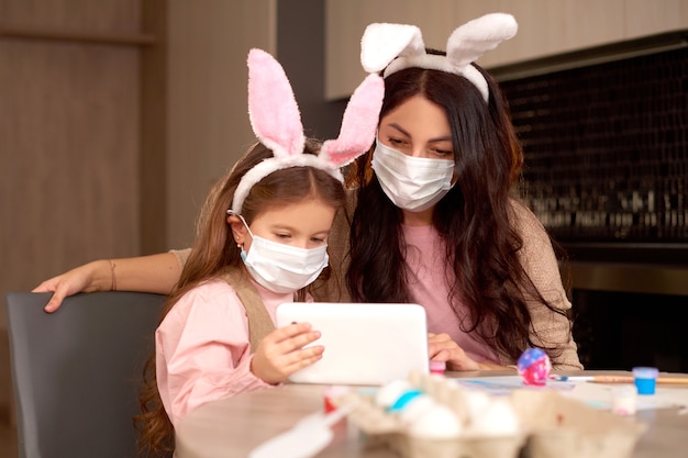 Figlia e madre in mascherina medica inviano videomessaggio ai parenti utilizzando un tablet. la famiglia festeggia la Pasqua a casa in quarantena online