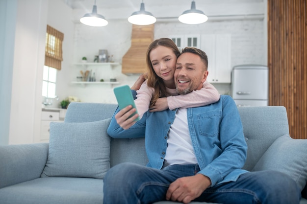 Figlia felice che abbraccia il collo del padre, che è seduto sul divano, si fotografa con suo padre su uno smartphone.