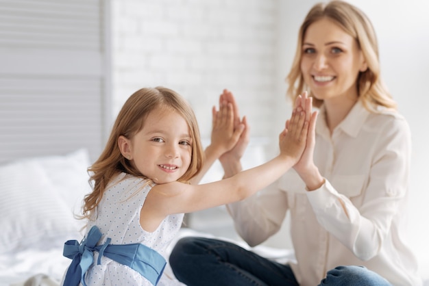 Figlia graziosa che lega a sua madre premendo i suoi palmi contro i palmi di sua madre mentre guarda davanti con un ampio sorriso.