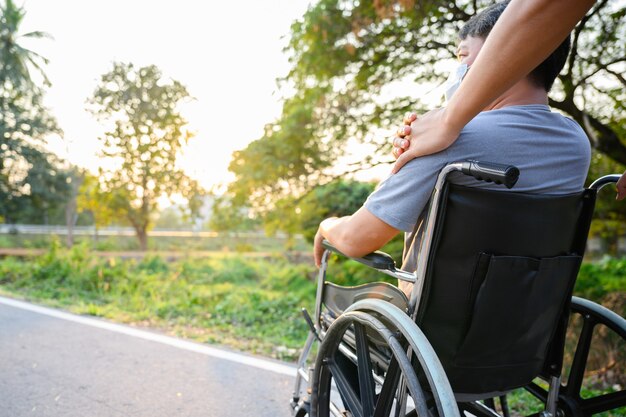 Figlio asiatico Camminando con il padre disabile in sedia a rotelle nel parco Aiutare il padre disabile spingendo una sedia a rotelle