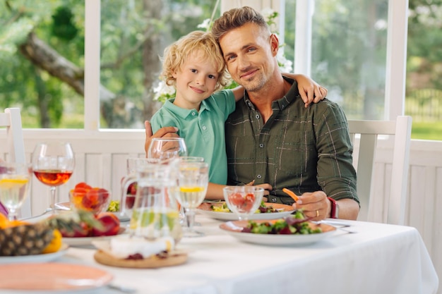 Figlio dagli occhi scuri che abbraccia il padre mentre consuma un pranzo delizioso