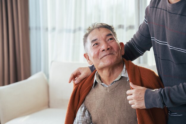 Figlio felice che copre suo padre con una coperta nella loro casa. Uomo anziano e famiglia felice.