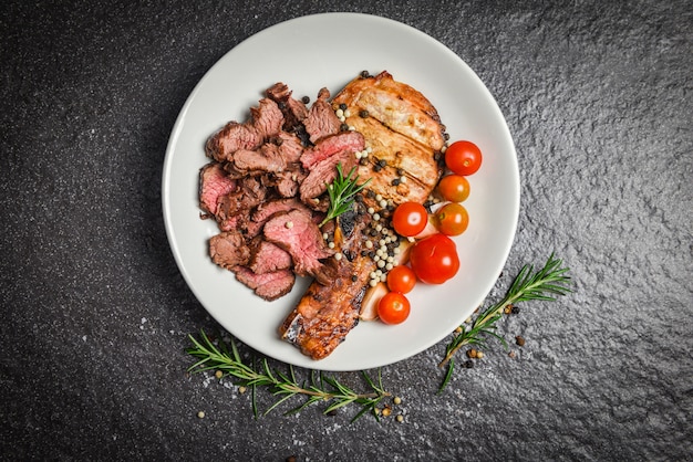 Filetto di manzo arrosto e braciole di maiale con erbe e spezie servire con verdure sul piatto bianco - fetta di carne di manzo alla griglia su sfondo nero