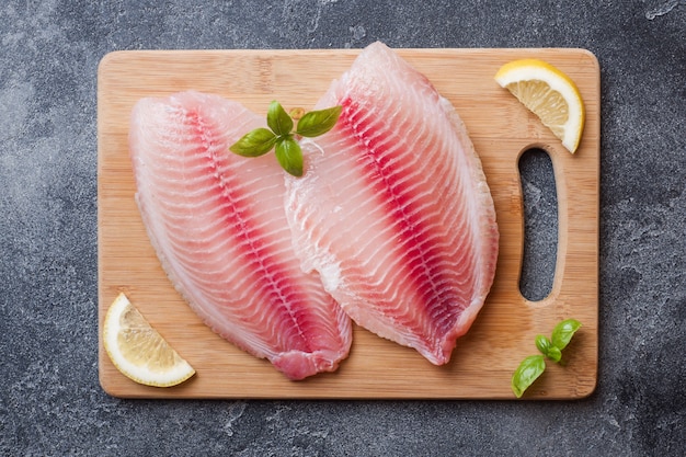 Filetto di pesce crudo di tilapia su un tagliere con limone e spezie. Tavolo scuro