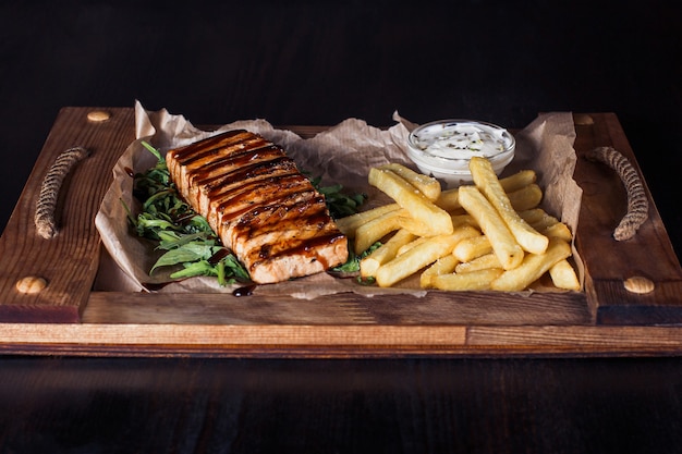 Filetto di salmone con patatine fritte su un vassoio di legno, bella porzione, superficie scura.