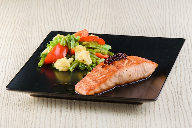 Filetto di salmone con verdure fritte su piatti quadrati neri su un tavolo di legno bianco.