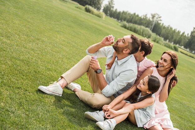 Fine settimana perfetto famiglia felice seduta sull'erba nel parco e soffiando bolle di sapone