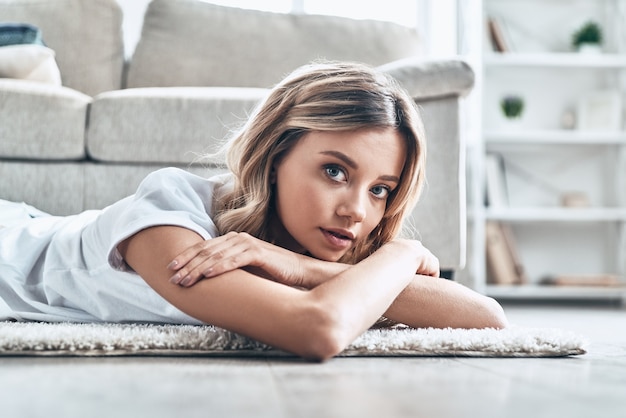 Fine settimana pigro. Bella giovane donna che guarda la macchina fotografica e sorride mentre è sdraiata sul pavimento a casa