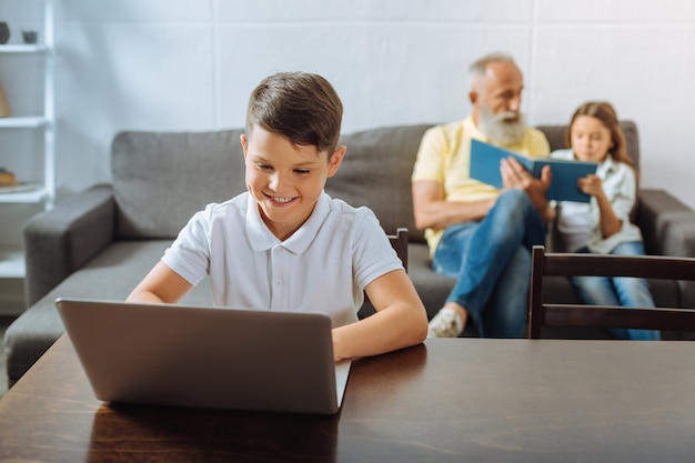 Fine settimana rilassato. Piacevole ragazzo carino che gioca un gioco sul computer portatile mentre suo nonno e la sorellina leggono un libro blu insieme sul divano