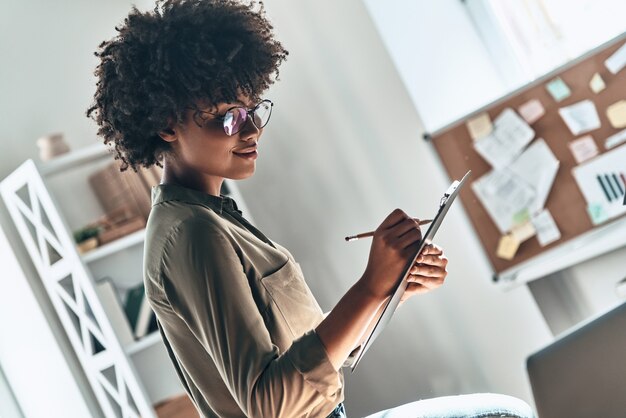 Finire di fare le cose. Attraente giovane donna africana che scrive qualcosa mentre lavora in ufficio