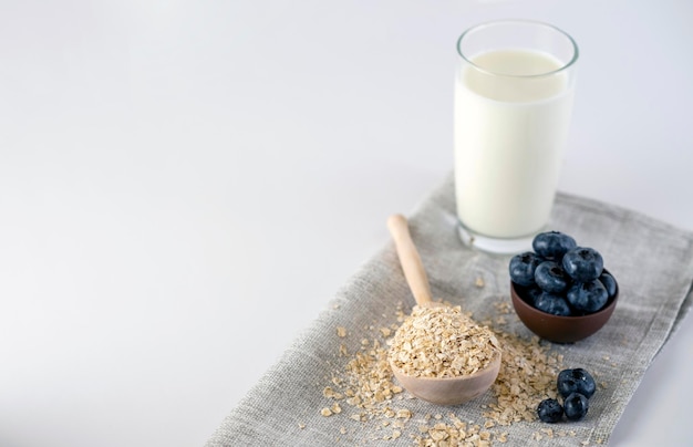 Fiocchi di avena cruda con mirtilli neri e un bicchiere di latte Angolo alto di mirtilli freschi e un cucchiaio di legno con farina d'avena posta sul tavolo vicino a un bicchiere di latte e su sfondo bianco in una stanza luminosa