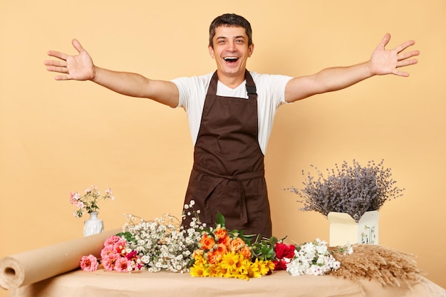 Fioraio uomo eccitato che fa bouquet in un negozio di fiori in piedi isolato su sfondo beige che allarga le mani facendo un gesto di benvenuto che invita al suo negozio di fiori