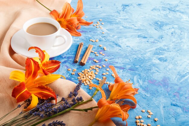 Fiori arancio della lavanda e del giglio di giorno e una tazza di caffè su un fondo concreto blu, con il tessuto arancio. vista laterale.